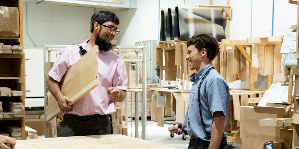 Two workers in a woodshop smiling and talking, one holding a wooden panel.
