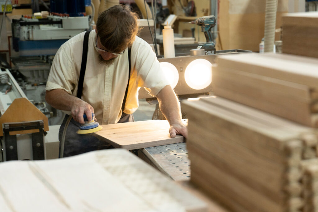 A woodworker wearing suspenders sanding a wooden plank in a workshop illuminated by bright lights.