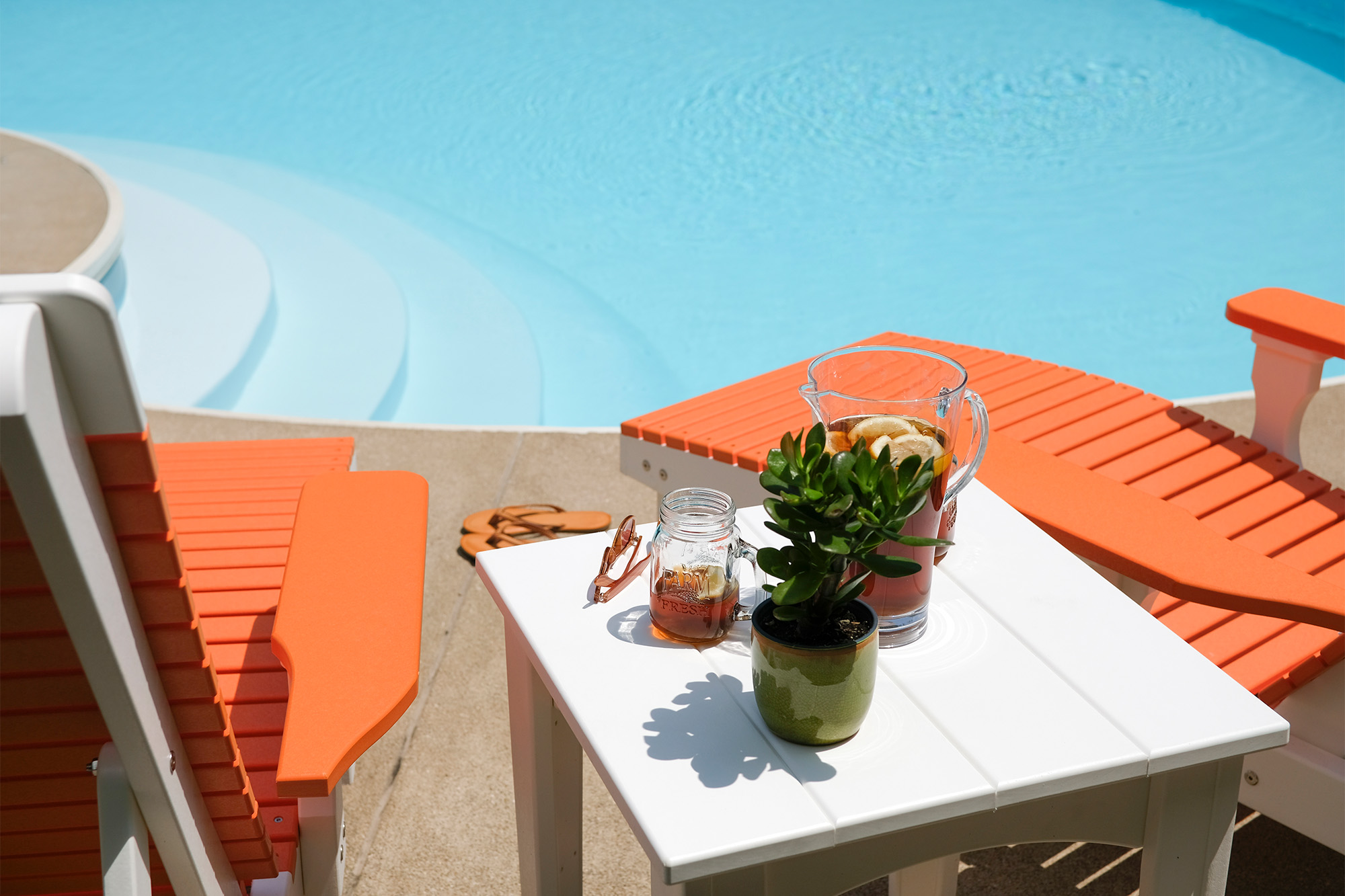 Two LuxCraft Furniture Adirondack chairs with bright orange accents, positioned by a pool, with a white side table holding a pitcher of iced tea, a mason jar glass, and a small potted plant.