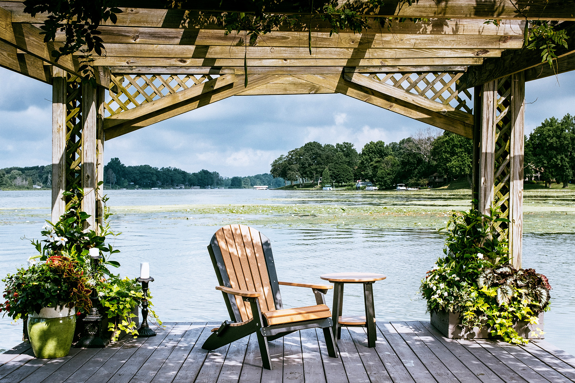 A serene lakeside setting featuring a LuxCraft Furniture folding Adirondack chair in cedar and black, positioned under a wooden pergola. The chair is accompanied by a matching side table, with lush greenery and water in the background.