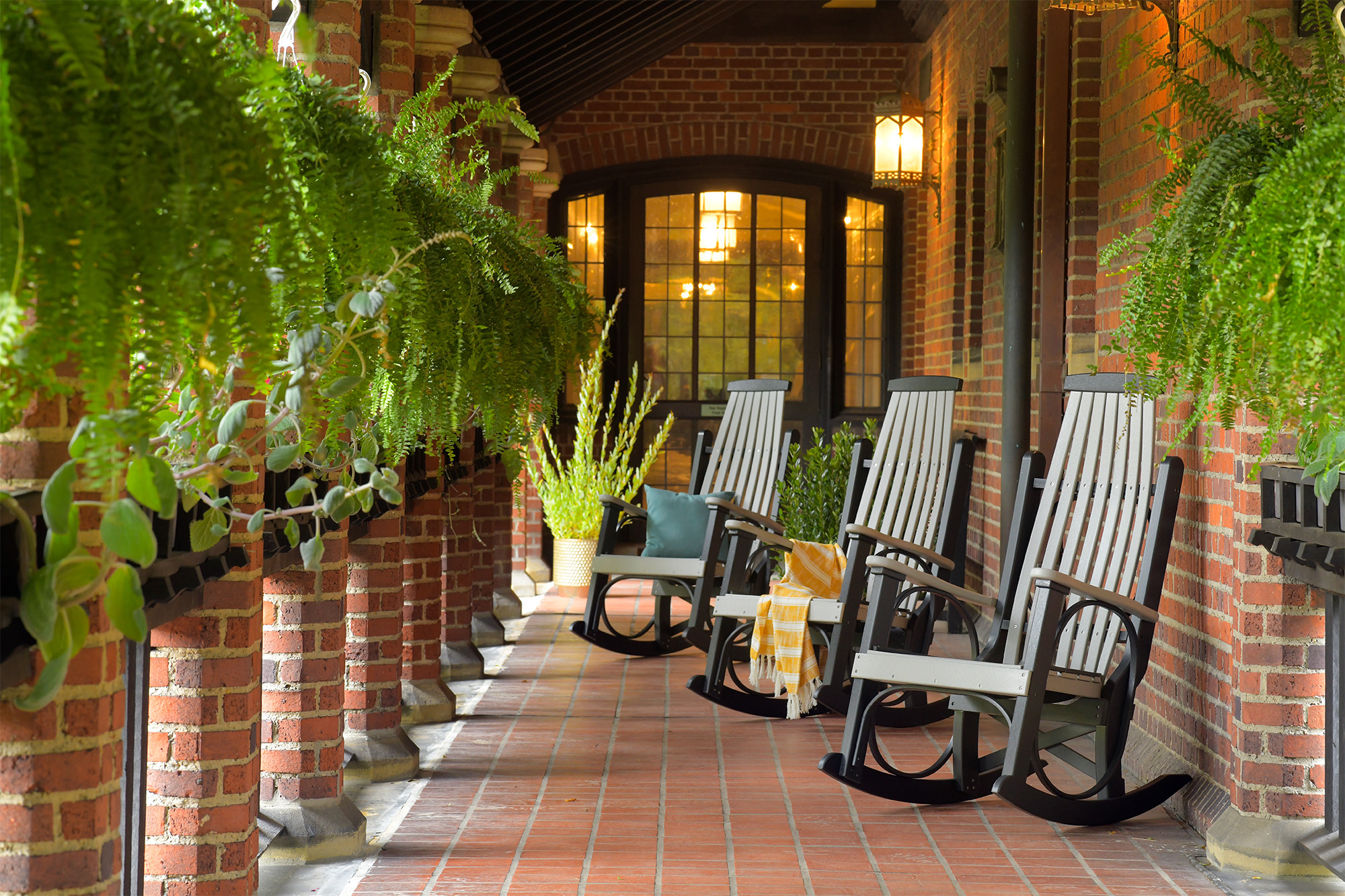 A cozy porch setting featuring LuxCraft Furniture’s Grandpa’s Rockers in dove gray and black. The rockers are arranged along a brick wall with lush green ferns hanging above, creating an inviting atmosphere.