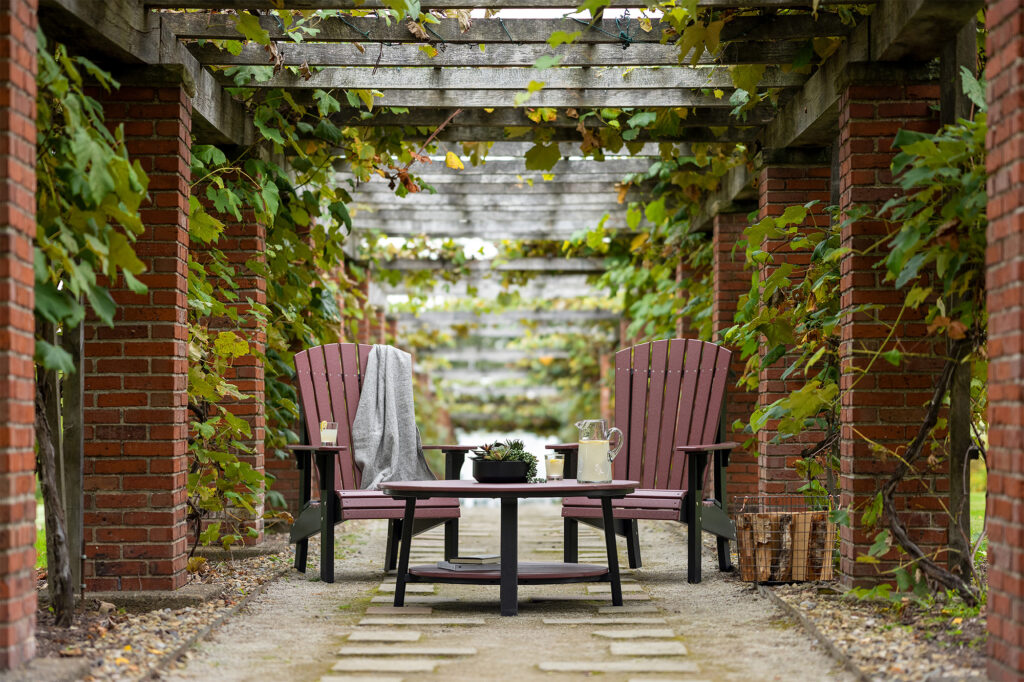 A pair of LuxCraft Furniture Royal Adirondack chairs in cherrywood and black finish, set under a pergola surrounded by greenery. A small table between the chairs holds a pitcher and a plant, creating a peaceful outdoor seating area.