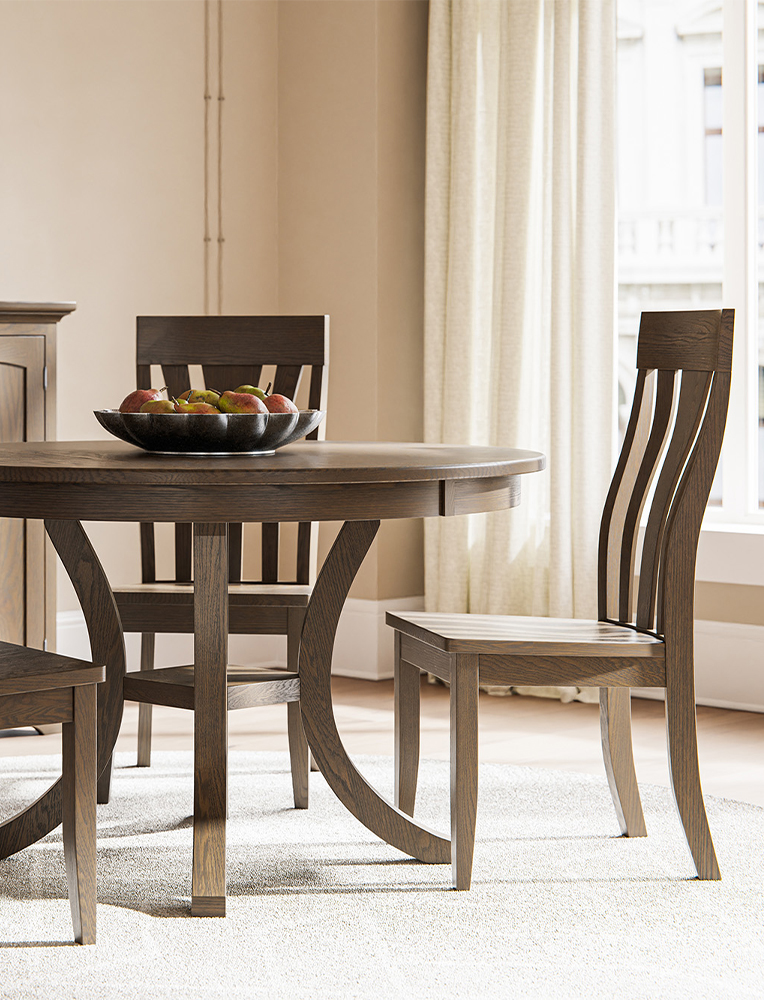 A sleek Amish dining set featuring a round table with curved wooden legs and matching slat-back chairs in a warm antique slate finish. The minimalist setting highlights the elegant craftsmanship available at Amish stores in Hamptonville, NC.