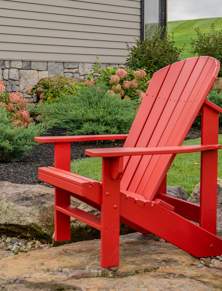 A vibrant red Amish Adirondack chair placed on a stone patio in a beautifully landscaped backyard, surrounded by greenery and blooming flowers. A striking example of durable and stylish Amish outdoor furniture.