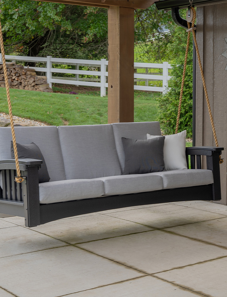 A handcrafted Amish outdoor porch swing with a sleek black frame, comfortable gray cushions, and sturdy rope hangers, set in a serene backyard with lush greenery and a white fence. A perfect example of durable and elegant Amish outdoor furniture.