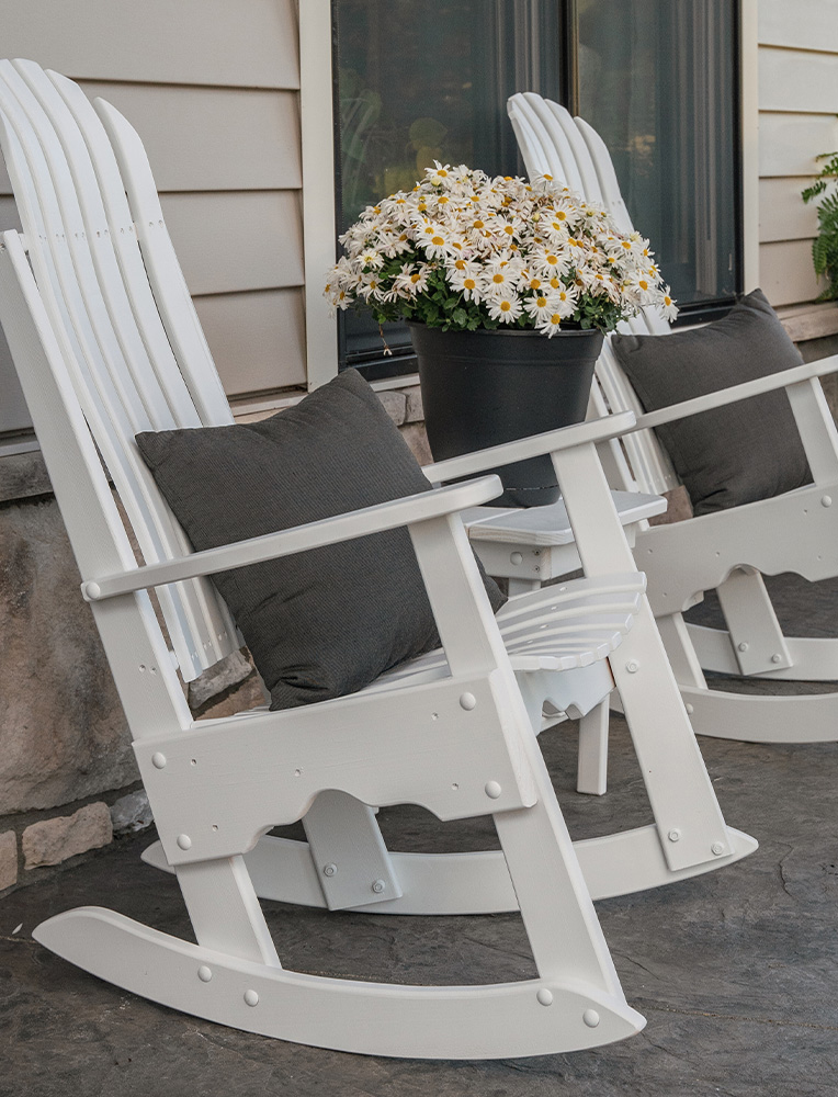 A pair of white Amish rocking chairs with gray cushions, placed on a stone patio next to a pot of blooming daisies. A classic and comfortable example of handcrafted Amish outdoor furniture.